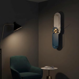 Op de afbeelding: Een moderne wandklok met een blauw en wit ovaal ontwerp. De klok heeft goudkleurige wijzers en een cirkelvormig midden. De klok is bevestigd aan een donkergrijze muur, met een goudkleurig frame. De klok is ongeveer 91 cm hoog.