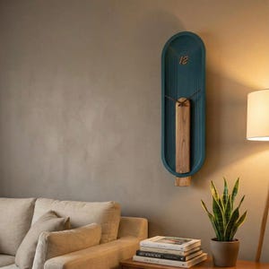 May include: A teal wall clock with a wooden feature. The clock has a long, oval shape with the number 12 at the top. The clock hands are thin and brown. The clock is mounted on a neutral-coloured wall.
