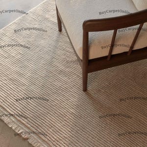 Könnte beinhalten: Ein beigefarbener, strukturierter Teppich mit einem linearen Muster. Der Teppich besteht aus einem gewebten Material und hat einen Fransenbesatz an einer Kante. Ein Holzarmlehnstuhl mit cremefarbenem Kissen ist teilweise zu sehen, was auf ein Wohnzimmer oder eine Wohndekoration hindeutet.