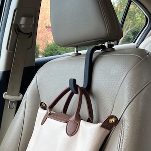 May include: A black car headrest hook holding a white and brown handbag. The handbag has brown handles and trim, with gold-colored hardware. The car seat is beige, and the background shows a glimpse of trees and a building.