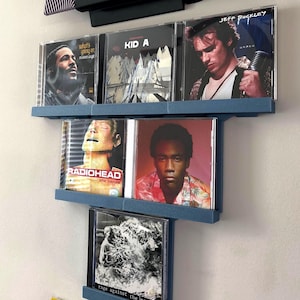 May include: A wall-mounted display shelf holding several CD cases, including albums by Jeff Buckley, Radiohead, and Rage Against the Machine. A vinyl record is displayed above the CDs. The shelf is painted blue.