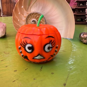 May include: An orange, round Halloween pumpkin container with a green stem and a cartoon face. The face has large white eyes, long black eyelashes, and a white mouth. The container is on a green surface.