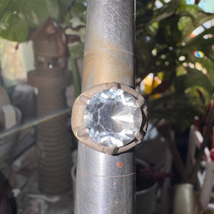 May include: A ring featuring a large, round, clear faceted gemstone set in a dull, matte-finish metal band. The ring is positioned on a tapered, cylindrical metal mandrel used for sizing, which shows horizontal markings along its length.
