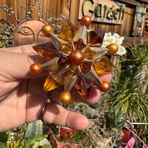 May include: A starburst-shaped decorative object with amber and gold tones. The object features a central cluster of amber-colored, faceted elements surrounded by gold-colored, pointed shapes and smaller gold spheres. The object is held in a hand against a blurred outdoor background.