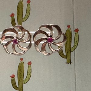 May include: A pair of silver-toned metal earrings featuring a swirling, pinwheel-like design with open cutouts. Each earring is centered with a small, round, vibrant pink gemstone. The earrings are displayed on a clear, curved plastic stand.