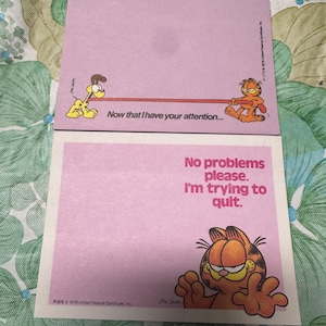 May include: Two pink Garfield notepads. One notepad has the text "Now that I have your attention..." with Garfield and Odie. The other notepad has the text "No problems please. I'm trying to quit." with Garfield.