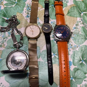 May include: A collection of vintage timepieces, including a pocket watch with a chain, a gold-toned wristwatch with a mesh band, a black leather strap watch, and a watch with an orange leather strap. The watches have various face designs.