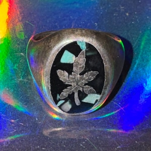 May include: A silver-toned ring featuring an oval-shaped inlay of black material accented with small, light blue stone fragments. The center of the inlay displays a raised, detailed silver-toned maple leaf design with visible veins and serrated edges.