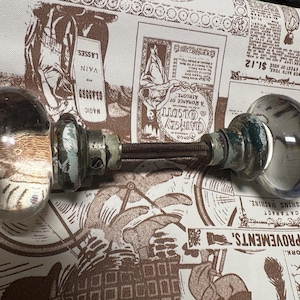 May include: A pair of vintage glass doorknobs with metal hardware. The clear glass knobs have a rounded shape and are connected by a long, threaded rod. The metal components show signs of age and wear. The background features a patterned paper with vintage advertisements.