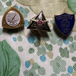 May include: Three vintage pins. One gold-toned pin with a clock face and the text "INSTRUMENT SOCIETY OF AMERICA". A black and gold triangle pin with a pink gem. A blue shield pin with "ENGINEERS & ARCHITECTS ASS'N. 1894".