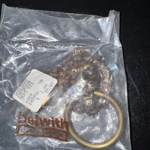 May include: A brass-toned metal lion head door knocker with a circular ring, enclosed in a clear plastic bag. The packaging features a white price tag with text including BUILDERS EMPORIUM and a brown logo with the text Belwith MADE IN USA.