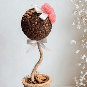 Könnte beinhalten: Ein dekoratives Kaffee-Bohnen-Topiary in einem Jute-Topf. Die kugelförmige Spitze ist mit Kaffeebohnen bedeckt, mit weißen Bändern und rosa Stoffblumen verziert. Der Topf enthält Zimtstangen und ist mit Bindfaden gebunden.