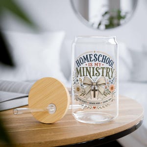 May include: A clear glass can-shaped tumbler with a bamboo lid and glass straw. The design features the text "HOMESCHOOL IS MY MINISTRY" in blue and green, a large beige bow with a cross, and floral accents with the text "RAISING HEARTS GROWING MINDS SERVING CHRIST".