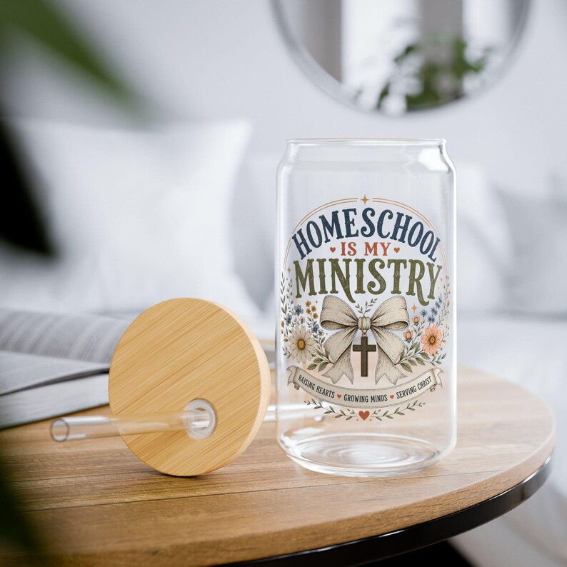 May include: A clear glass can-shaped tumbler with a bamboo lid and glass straw. The design features the text "HOMESCHOOL IS MY MINISTRY" in blue and green, a large beige bow with a cross, and floral accents with the text "RAISING HEARTS GROWING MINDS SERVING CHRIST".