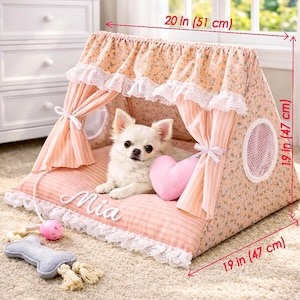 May include: A peach-colored pet tent with a floral print, white lace trim, and striped curtains. The base features the name Mia in white script. It measures 20 inches wide, 19 inches high, and 19 inches deep, with a small dog and toy inside.