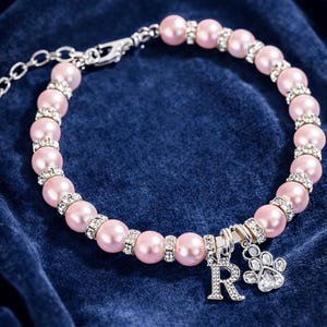May include: Pink pearl pet collar with silver accents. The collar features a series of pink pearls, interspersed with silver rhinestone spacers. A silver charm with the letter "R" and a paw print hangs from the collar. The collar is set against a dark blue background.
