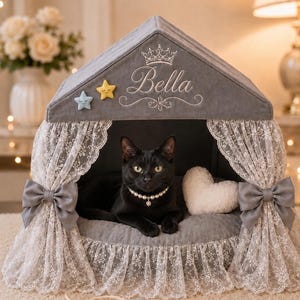 Pode incluir: Uma casa para animais de estimação cinzenta com telhado pontiagudo, com o nome bordado Bella, um ícone de coroa e duas pequenas aplicações de estrelas. A entrada tem cortinas de renda branca com laços cinzentos, uma almofada de coração e um gato.