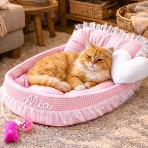 May include: A ginger cat rests in a pink and white gingham pet bed with a white heart-shaped pillow. The bed has a frilly trim and the name "Mia" is written on the side. A pink cat toy is nearby.