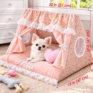 Puede incluir: Una cama para perros rosa floral con ribete de encaje blanco, que mide 51 cm de ancho y 47 cm de alto y de profundidad. Un perro blanco pequeño descansa dentro, con una almohada en forma de corazón rosa. La cama tiene un diseño tipo tienda.