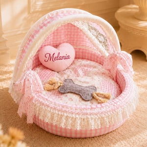 May include: A round, pink and white gingham pet bed with a canopy, featuring white lace trim and pink bows. Inside, a heart-shaped pillow is embroidered with the name Melania, accompanied by a grey felt bone toy with rope ends on a floral cushion.
