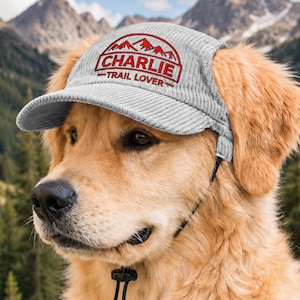 Op de afbeelding: Een lichtgrijze corduroy huisdierenpet met een geborduurde rode berggrafiek en de tekst CHARLIE TRAIL LOVER. De pet heeft uitsparingen voor de oren en een zwarte verstelbare kinband met een plastic koordstopper voor een goede pasvorm.
