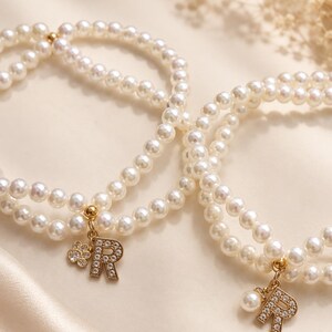 May include: Two double-strand bracelets made of white faux pearls. Each bracelet features a gold-toned charm in the shape of the letter R, encrusted with small clear crystals. One bracelet includes a small flower charm, the other a single pearl.