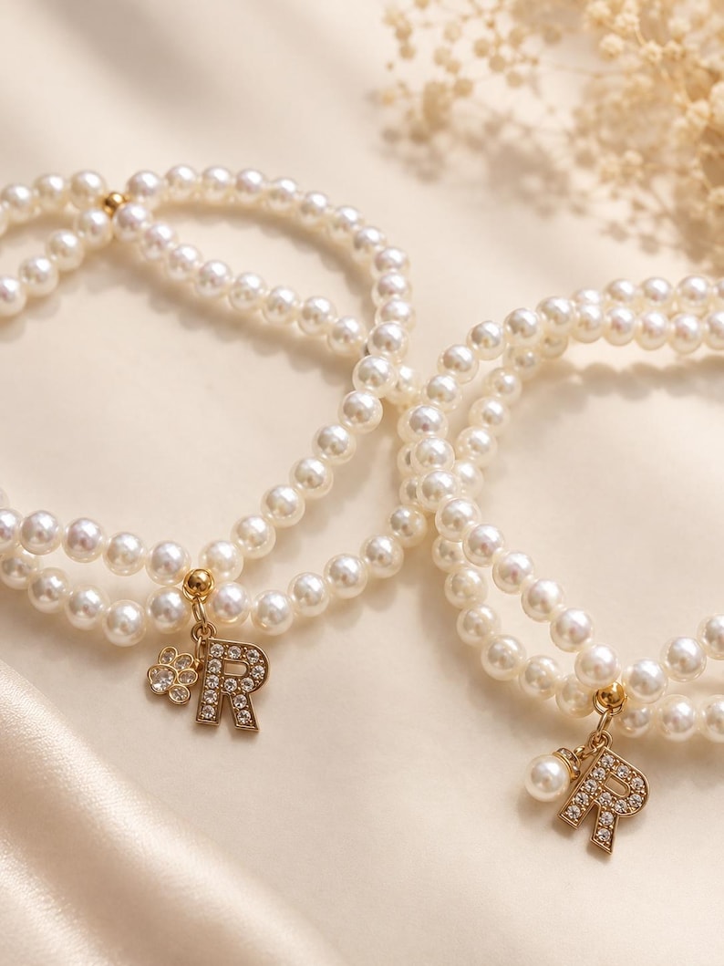 May include: Two double-strand bracelets made of white faux pearls. Each bracelet features a gold-toned charm in the shape of the letter R, encrusted with small clear crystals. One bracelet includes a small flower charm, the other a single pearl.