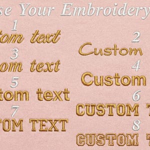 May include: A light pink fabric background displays eight numbered embroidery font options in gold thread. The text reads Choose Your Embroidery Font at the top, with a circular badge stating Professionally machine embroidered in the top right corner.