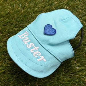 May include: A light blue pet cap featuring a curved brim with the name Buster embroidered in white thread. The crown of the cap is adorned with a small, dark blue embroidered heart patch. A thin black elastic strap is attached to the side.