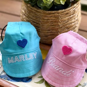 May include: Two pet hats, one light blue and one light pink, each featuring an embroidered heart and a name. The blue hat displays the name MARLEY in white block letters with a dark blue heart, while the pink hat shows Brandi in white script.