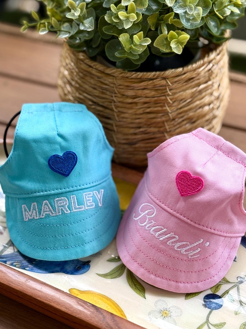 May include: Two pet hats, one light blue and one light pink, each featuring an embroidered heart and a name. The blue hat displays the name MARLEY in white block letters with a dark blue heart, while the pink hat shows Brandi in white script.