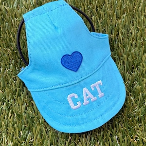 May include: A bright blue pet visor hat featuring a dark blue embroidered heart on the front panel and the word CAT stitched in white block letters on the brim. The hat includes a black elastic strap for securing it around the head.