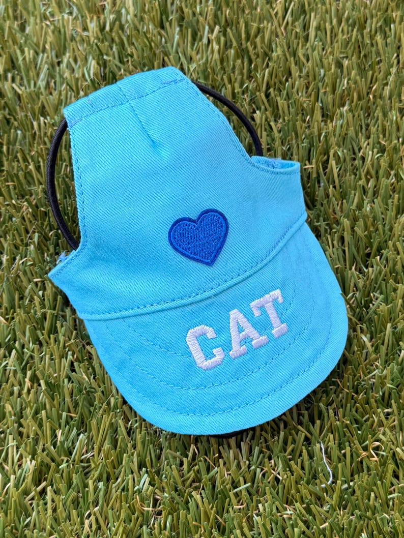 May include: A bright blue pet visor hat featuring a dark blue embroidered heart on the front panel and the word CAT stitched in white block letters on the brim. The hat includes a black elastic strap for securing it around the head.