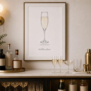 May include: A framed wall art print featuring a detailed illustration of a champagne flute filled with golden liquid. Below the glass, the text reads "CHAMPAGNE" in capital letters and "bubbles, please" in a cursive script font on a white background.