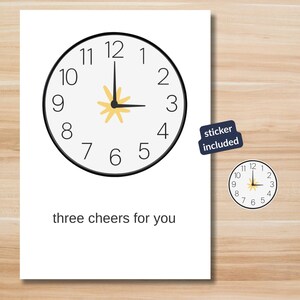 May include: A white greeting card with a black clock face showing 3 o'clock. The card has the text "three cheers for you" at the bottom. A small sticker with a clock face is included, and the words "sticker included" are printed on a blue sticker.