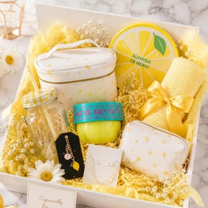May include: A gift box containing a white cosmetic bag with yellow lemon prints, a round yellow lemon-themed tin with the text SUMMER IS HERE, a yellow towel with a ribbon, a glass mason jar with a yellow lid, a body butter jar, and jewelry.