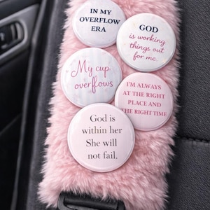 May include: Pink fluffy seatbelt cover with five round buttons. The buttons have inspirational quotes like "In my overflow era" and "God is working things out for me." The cover is attached to a black seatbelt.