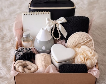 May include: A gift box filled with self-care items. Includes a black cosmetic bag, a white 2026 calendar, a gray and white essential oil diffuser, a pearl necklace, a heart-shaped box, and various spa accessories in neutral tones, all arranged in a brown box.