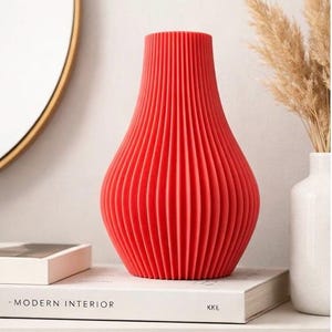 May include: A vibrant red decorative vase with a ribbed, textured surface. The vase has a bulbous shape and a narrow neck. It is displayed on a white shelf with a book that reads "- MODERN INTERIOR" and "KKE".