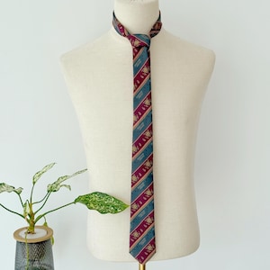 Puede incluir: Una corbata estampada exhibida en un maniquí. La corbata presenta rayas diagonales en verde azulado y burdeos, con un emblema y texto dorados repetidos. La corbata está anudada en el cuello, mostrando su diseño y combinación de colores.