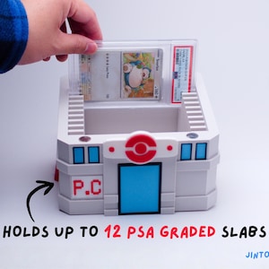 May include: A white and grey storage container with blue and red accents, designed to hold graded trading card slabs. The container has a red and white circular emblem and the text "P.C." on the side. The text "HOLDS UP TO 12 PSA GRADED SLABS" is also visible.