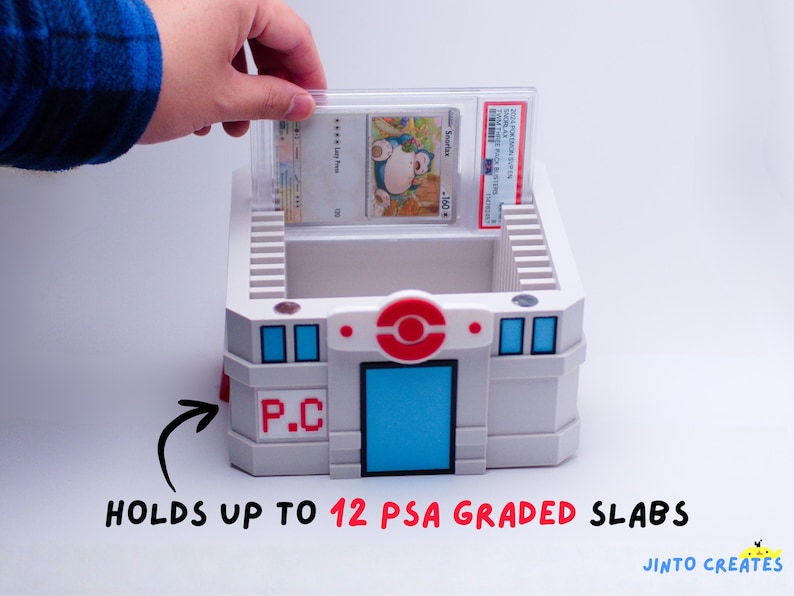 May include: A white and grey storage container with blue and red accents, designed to hold graded trading card slabs. The container has a red and white circular emblem and the text "P.C." on the side. The text "HOLDS UP TO 12 PSA GRADED SLABS" is also visible.