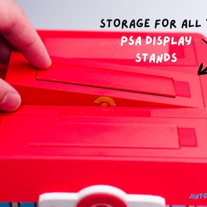 May include: A red plastic storage container with the text "STORAGE FOR ALL 3 PSA DISPLAY STANDS". The container has three angled slots for display stands. A hand is interacting with the container.