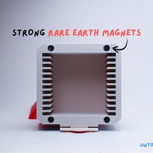 May include: A light grey rectangular object with vertical lines on the sides. The text "STRONG RARE EARTH MAGNETS" is written above the object. Four black magnets are visible on the corners of the object.