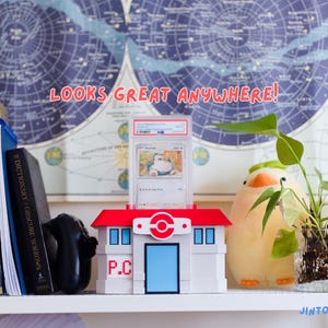 May include: A white and red Pokemon Center display case holding a graded Snorlax trading card. The case is on a shelf with books, a plush toy, and a plant. Text reads "LOOKS GREAT ANYWHERE!"