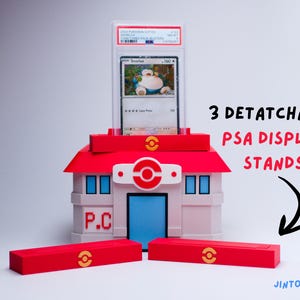 May include: A red, white, and blue building-shaped display stand for a graded trading card. The stand has a red roof and three detachable red bases. The card features a Snorlax Pok&eacute;mon. The stand is designed to hold the card.