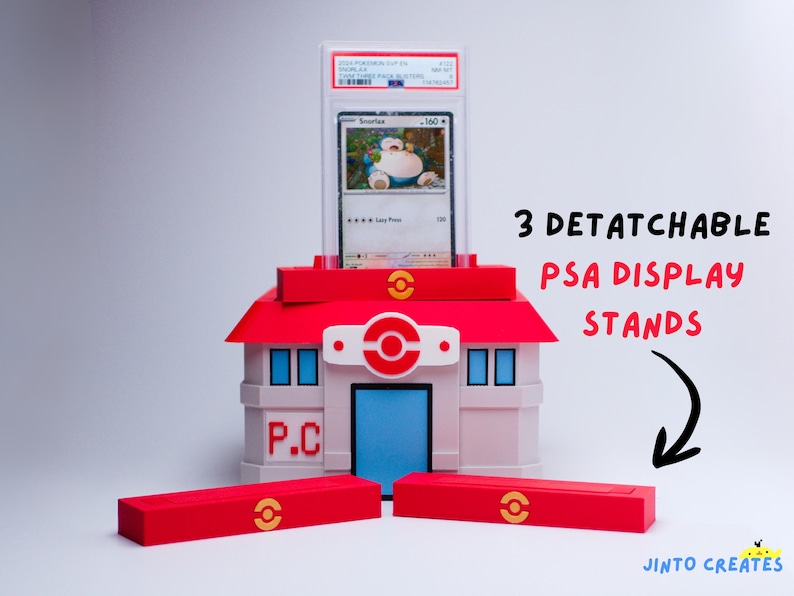 May include: A red, white, and blue building-shaped display stand for a graded trading card. The stand has a red roof and three detachable red bases. The card features a Snorlax Pok&eacute;mon. The stand is designed to hold the card.