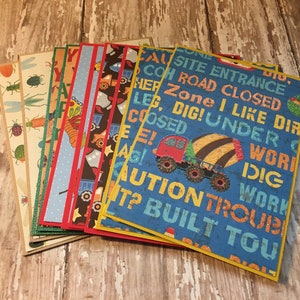 May include: A stack of eight colorful cards with construction vehicle designs and text. The top card is blue with yellow trim and features a yellow and black cement mixer with the text "Caution Trouble Built You" and "Site Entrance Road Closed Zone I Like Dirt Dig! Under Work Dig Work".
