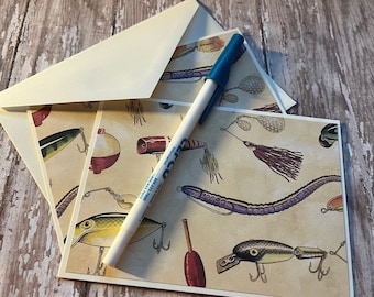 May include: Two cream-colored cards with a vintage illustration of fishing lures and a blue and white pen on top.