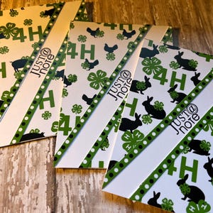 May include: Three white cards with a green and black 4-H clover pattern. Each card has a diagonal white stripe with black polka dots and the text "JUST@ note" in black. The cards are on a wooden surface.
